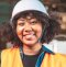 A woman with curly hair and a bright smile, wearing a white industrial safety helmet and a yellow safety gown.